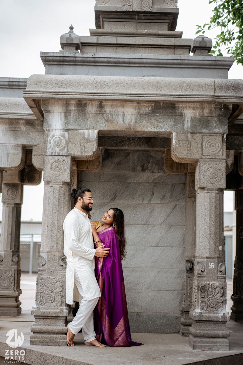 A couple poses romantically under ornate stone pillars at a temple, with one wearing white and the other in purple.
