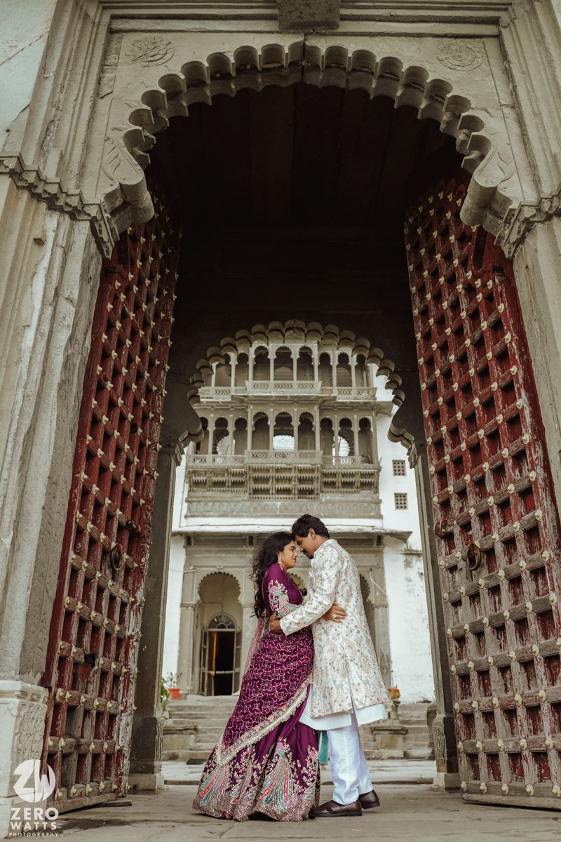 Traditional Indian couple in wedding attire embracing under ornate archway of historic palace architecture.