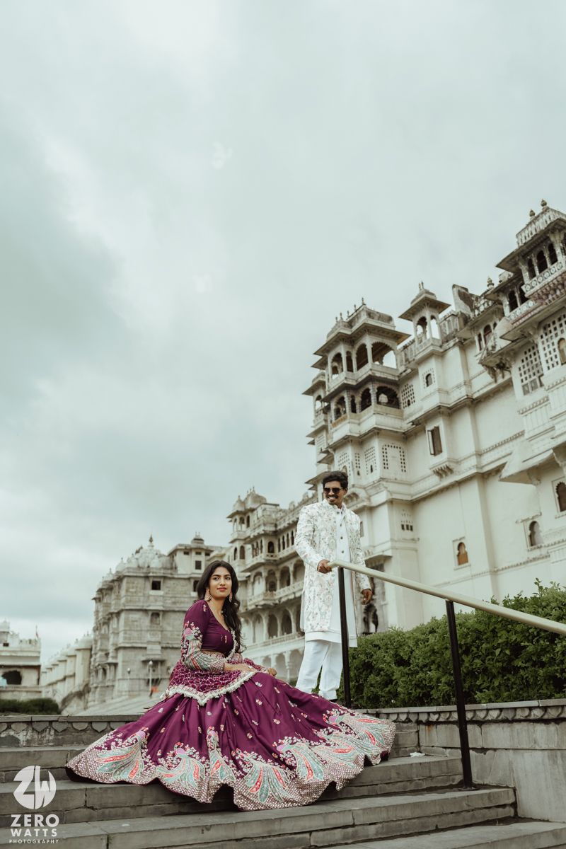 Traditional Indian couple poses dramatically on palace steps as woman twirls purple lehenga dress.