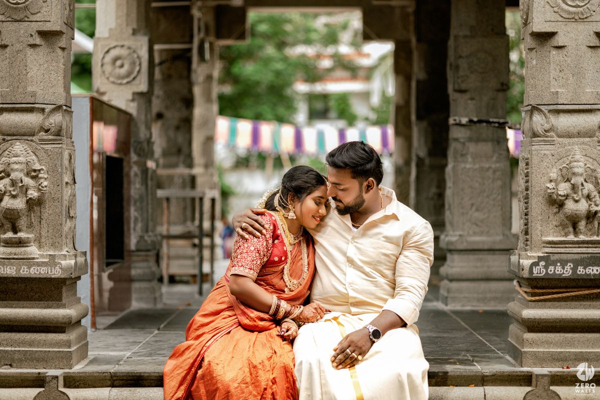 Romantic couple in orange sari and white kurta embracing in a traditional temple setting with ornate architecture.