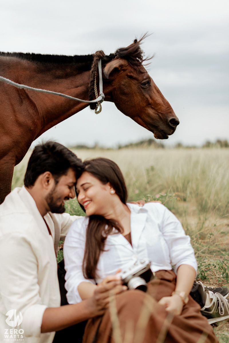 A romantic couple shares tender moments in a field with a brown horse in the background.