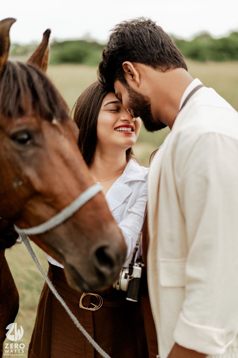 Romantic couple shares tender moments in a field with a brown horse beside them in neutral attire.