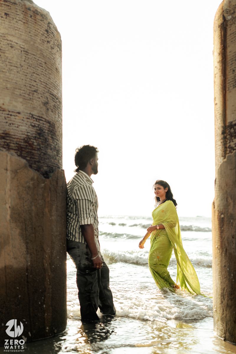 A couple poses playfully in shallow water against a brick wall during a romantic photoshoot.