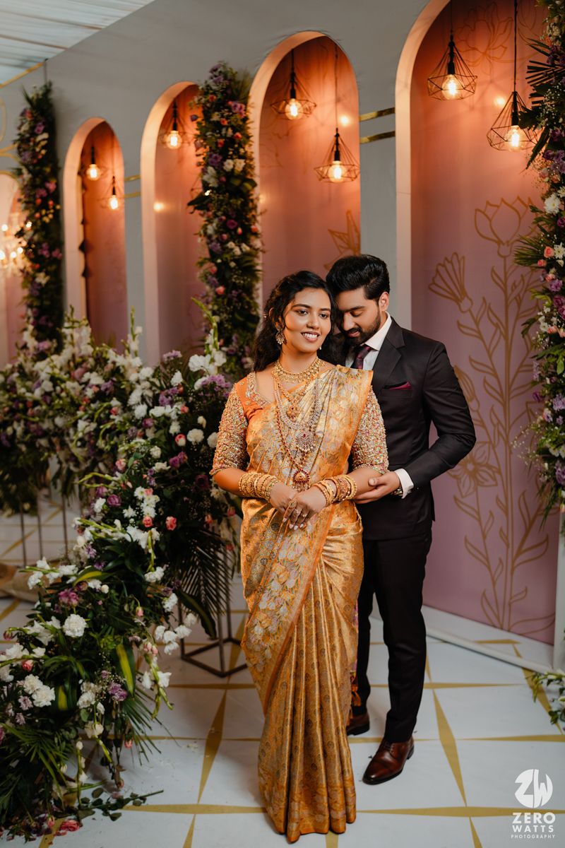 A couple poses in traditional formal attire against pink arched walls decorated with hanging lights and flowers.