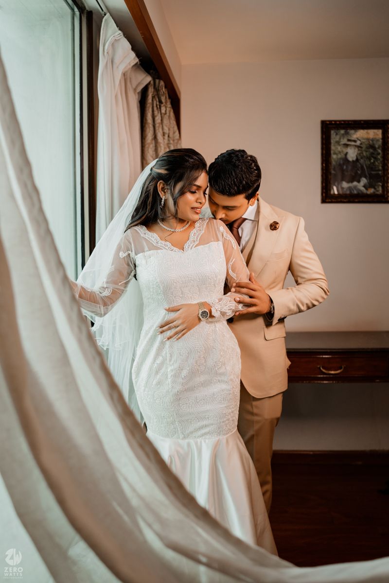 A romantic wedding moment captured through sheer curtains in a softly lit indoor setting.