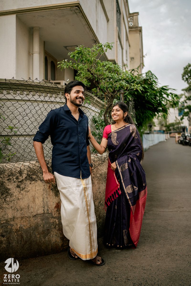 A couple in traditional South Indian attire share candid moments together against a building wall.