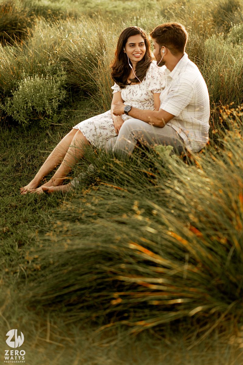 A romantic couple sits together in tall grass during a golden hour photoshoot wearing white outfits.