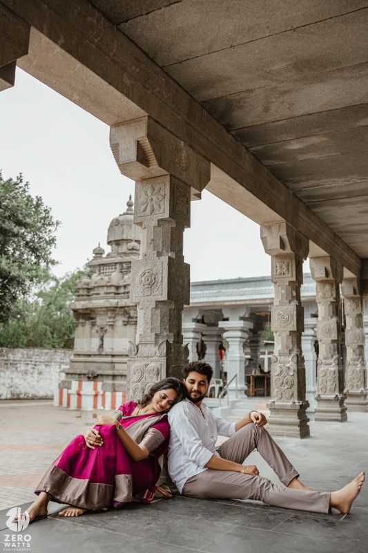 A couple relaxes on the ground beneath ornate stone pillars, with one wearing a vibrant pink outfit.