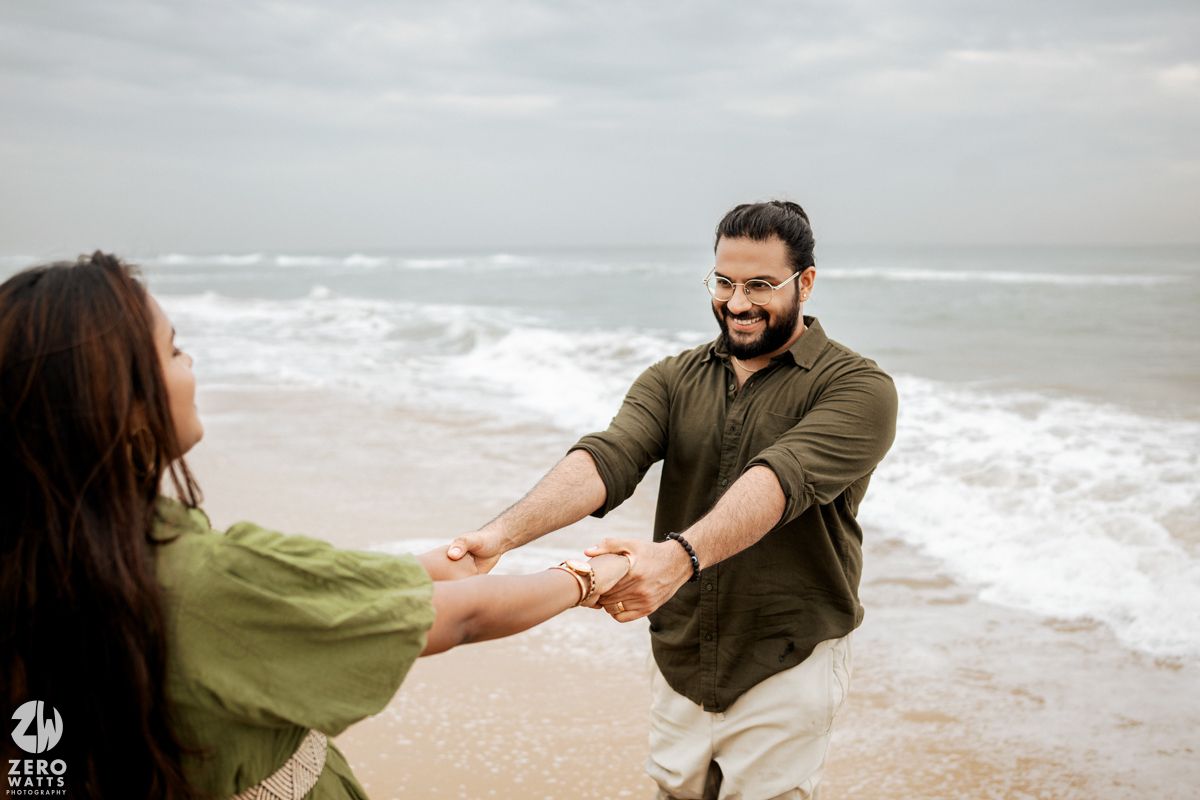 A couple in olive green outfits embraces on a misty oceanfront beach.