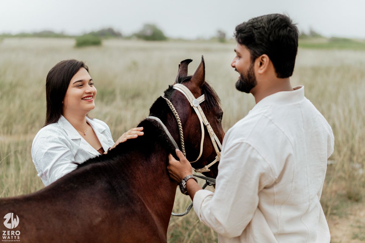 A couple shares a tender moment together in a field with horses visible behind them.