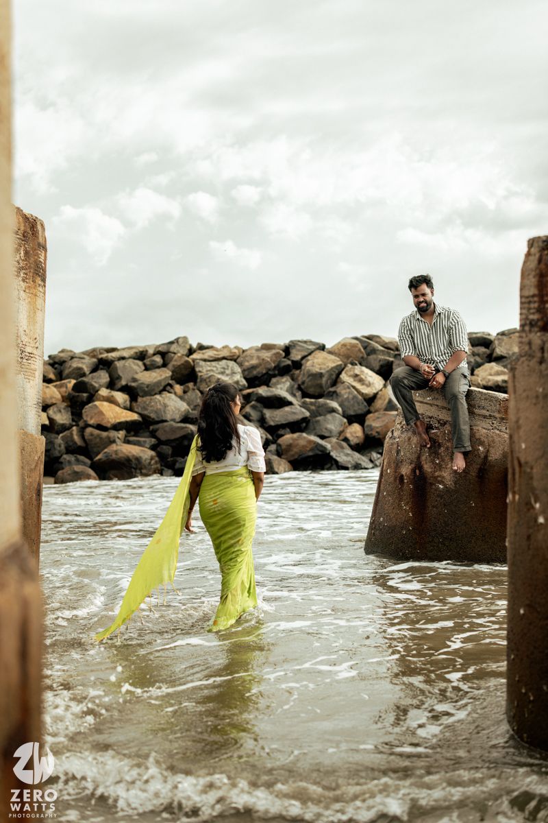 Artistic beach photoshoot with couple wading through water between concrete pillars.