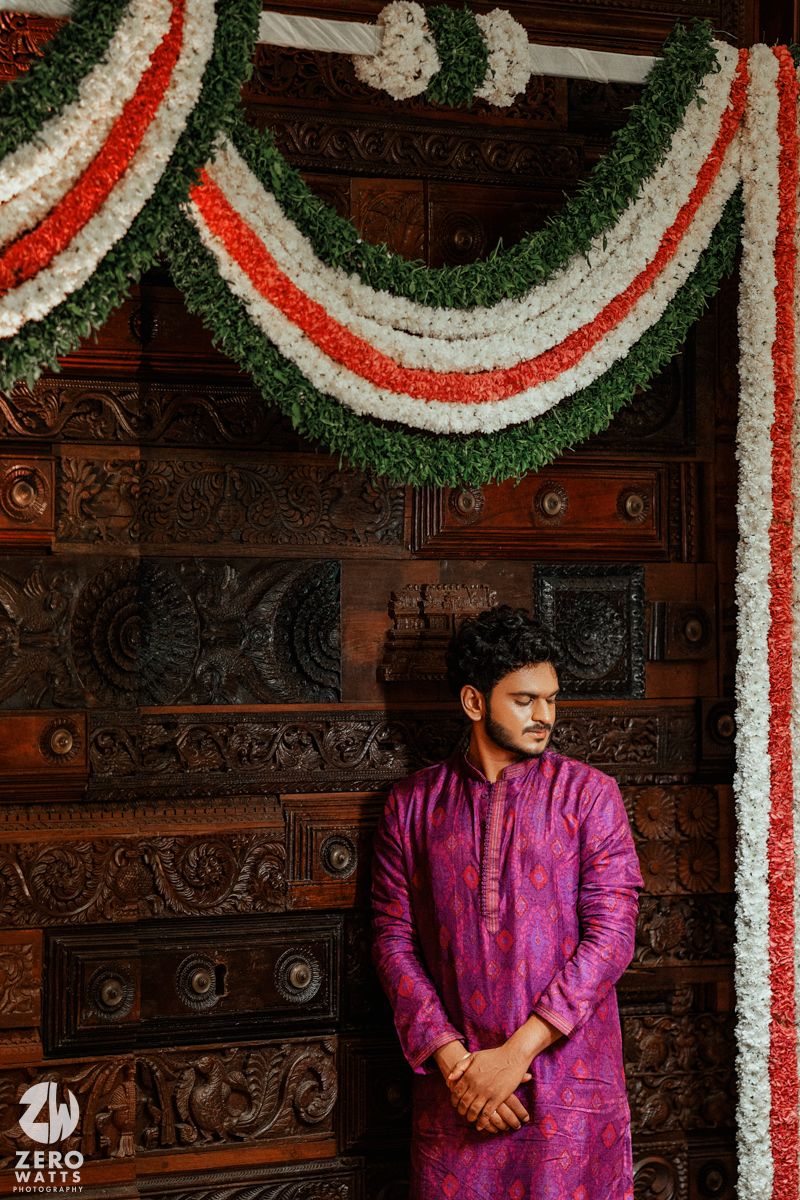 Young man in purple kurta stands before ornate wooden door adorned with tricolor flower garlands.