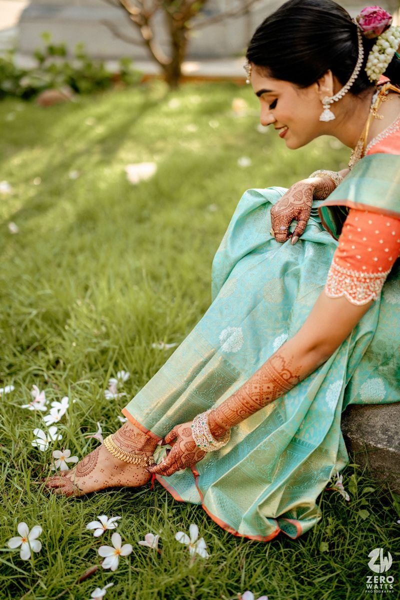 Indian bride in teal Kanjivaram saree with mehndi-adorned hands sits on grass adjusting gold anklets.