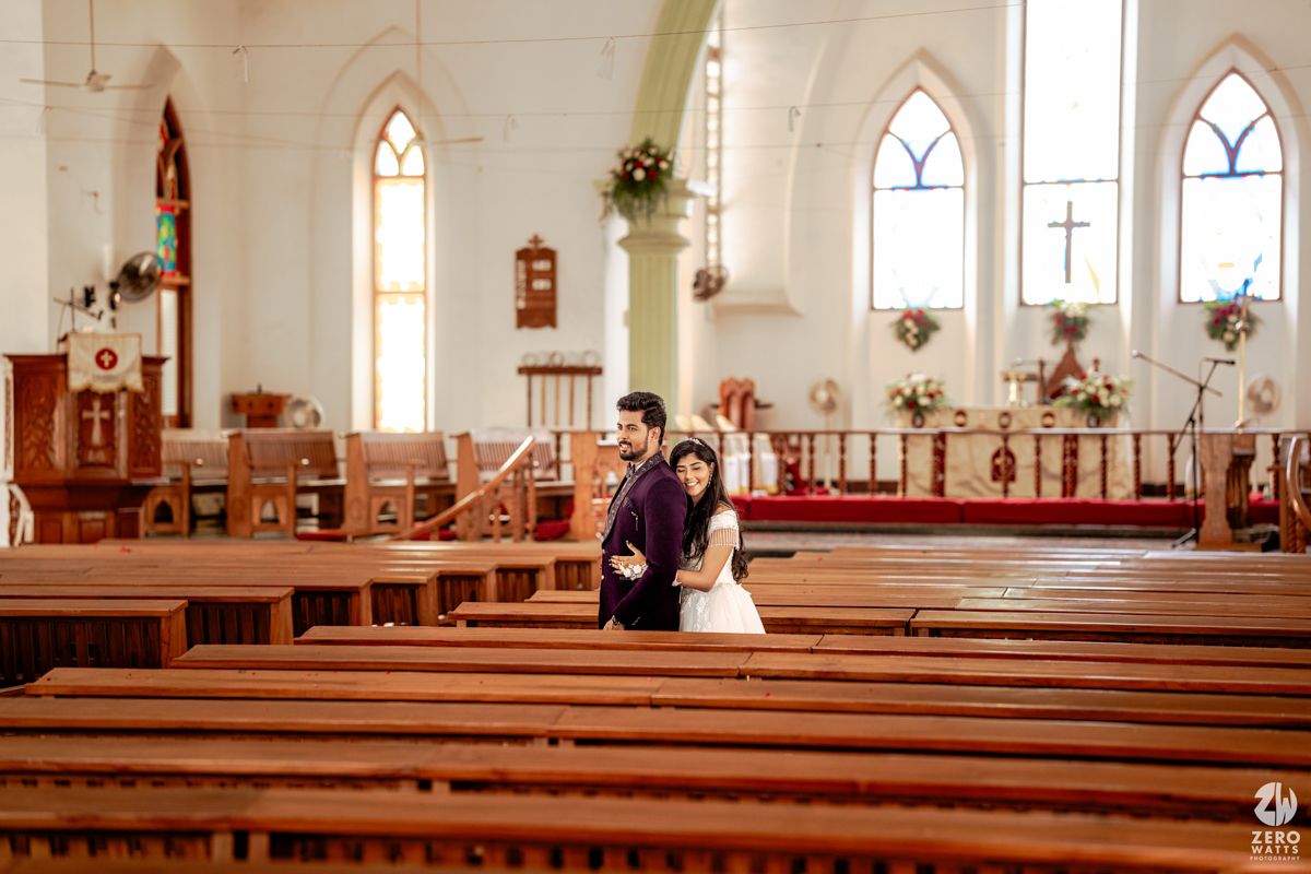 Couple poses romantically between wooden church pews in a bright white chapel with arched windows.