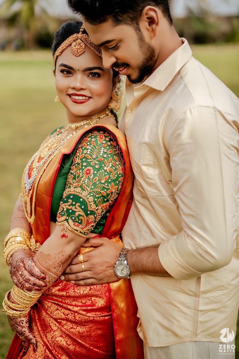 South Indian bride in red Kanjivaram saree with green blouse and gold jewelry poses with groom in cream shirt.