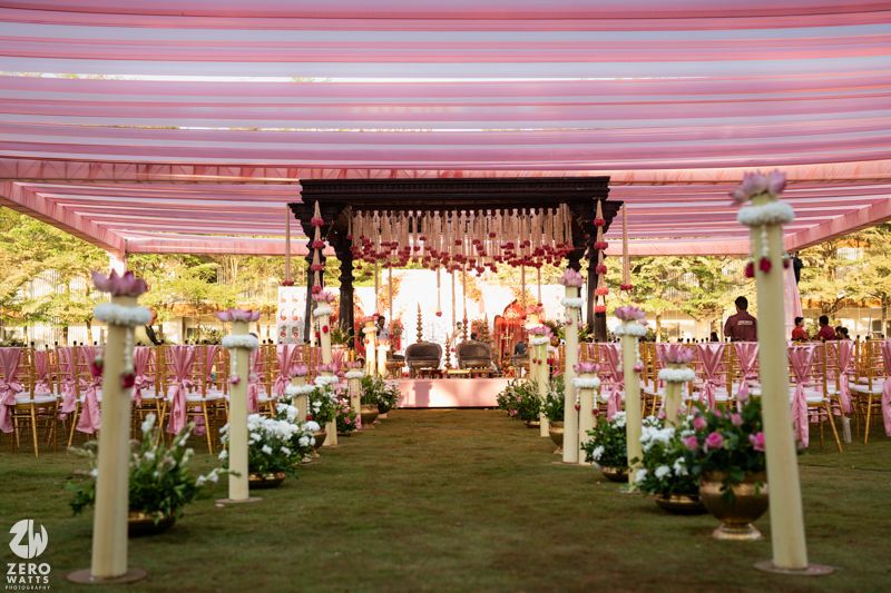 Elegant outdoor Indian wedding venue with floral aisle, pink canopy, decorated mandap stage, and rows of chairs at dusk.