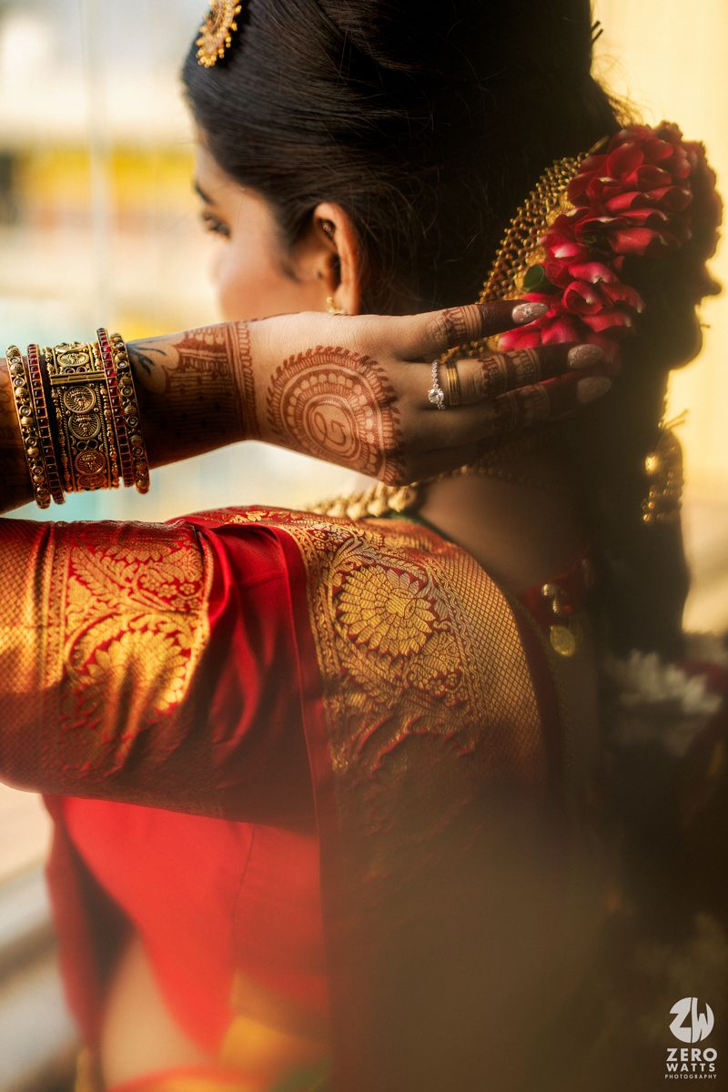 South Indian bride showcasing intricate mehndi henna designs on hands wearing red silk saree with gold bangles and floral hair accessories.
