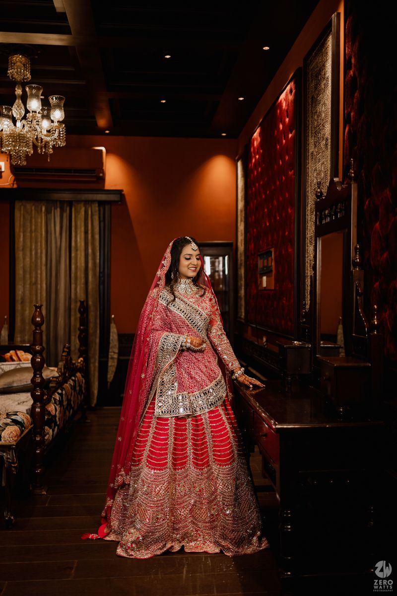 Traditional Indian bride wearing an ornate red and gold lehenga poses elegantly in a luxurious interior setting.