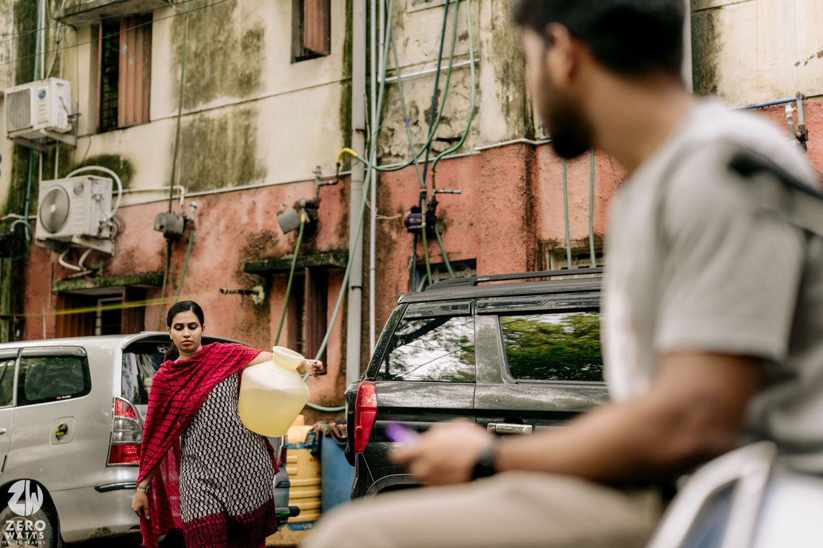 Urban street scene with people walking past weathered buildings and parked vehicles.
