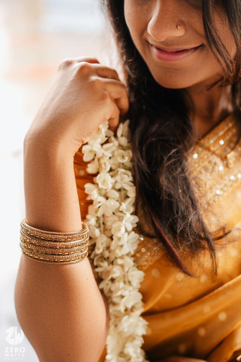 Close-up view of gold bangles and traditional jewelry on a woman's wrist with white jasmine flowers in soft lighting.