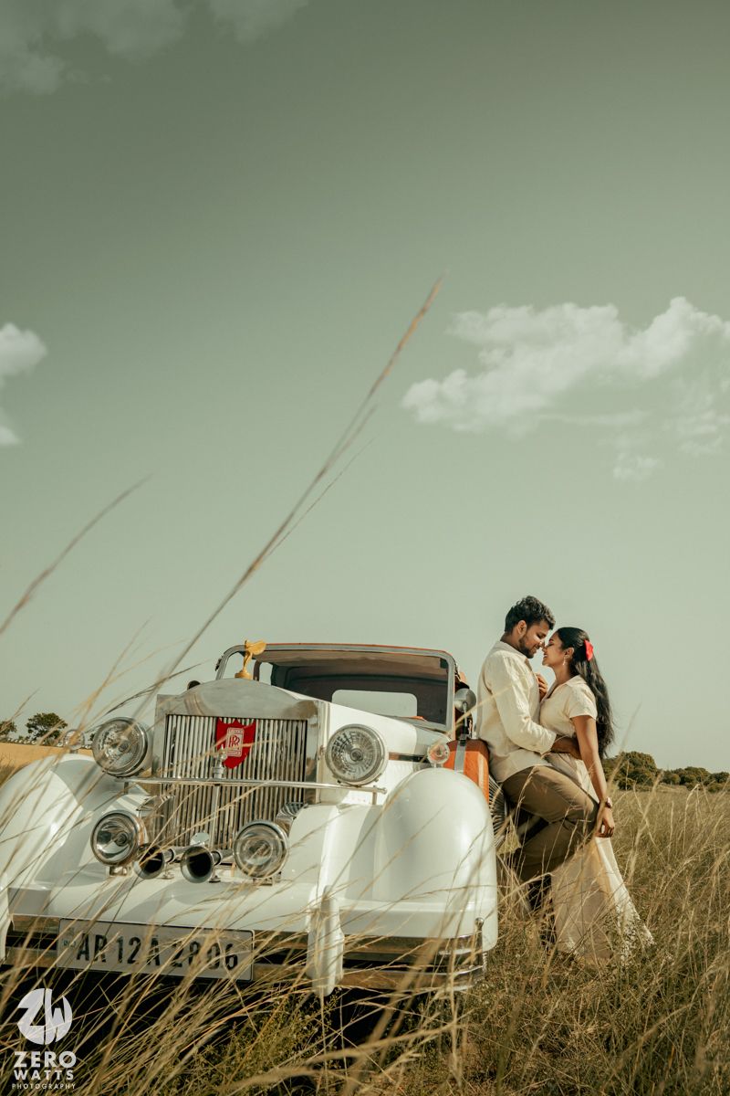 Couple sharing an intimate moment beside a classic white automobile in a field at sunset.