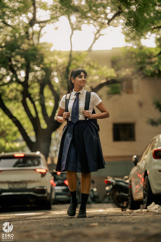 A student in a navy dress and backpack walks through a tree-lined campus path in warm afternoon sunlight.