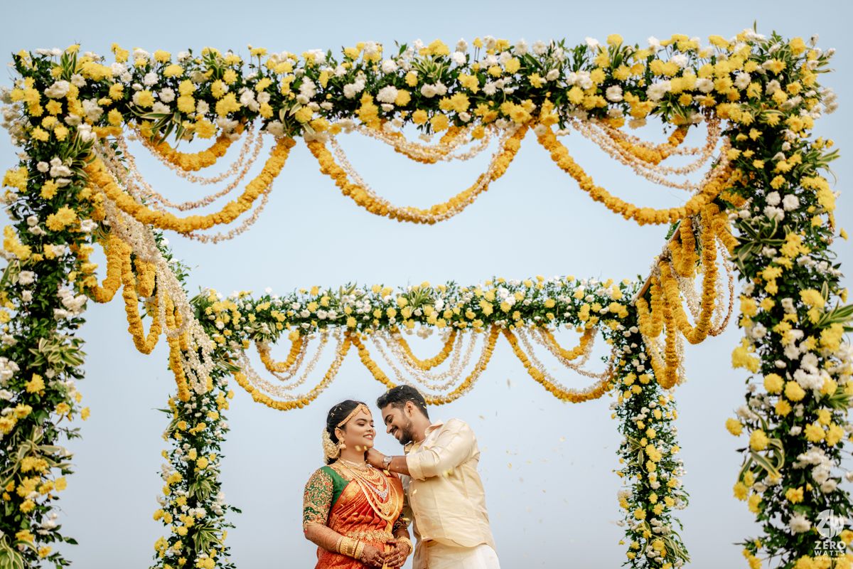 Indian couple sharing a romantic moment under a vibrant yellow floral wedding mandap arch.