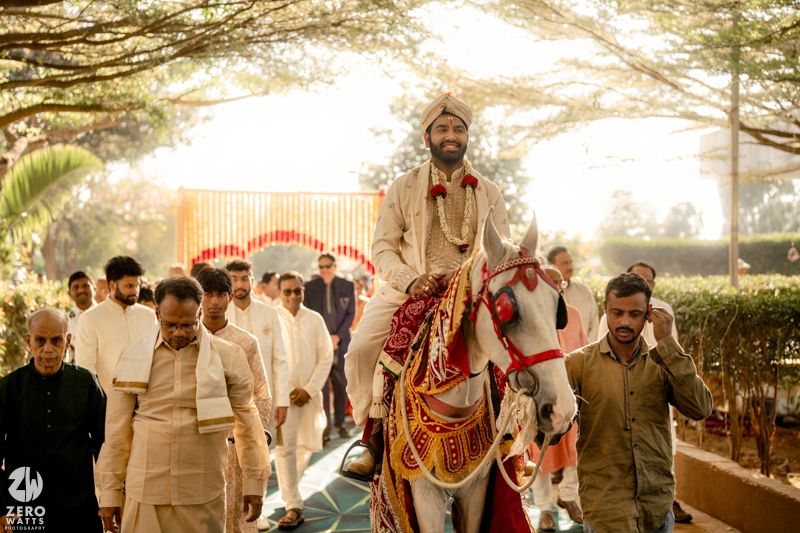 Indian groom in traditional attire rides a decorated white horse during a baraat wedding procession surrounded by guests.