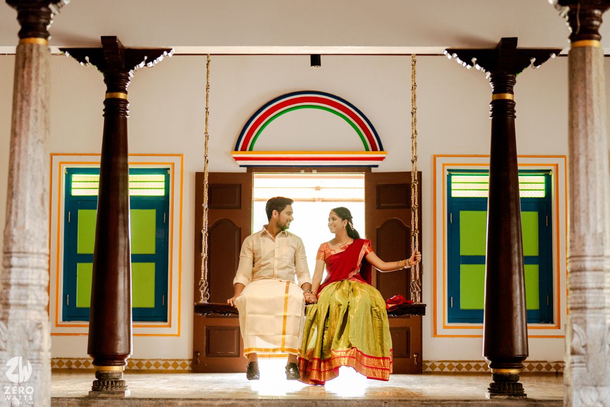 Couple in traditional South Indian attire sitting on a swing in a colorful heritage home with wooden pillars.