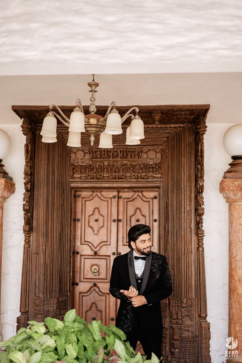 Groom in black tuxedo standing in front of ornate wooden door with decorative archway at elegant wedding venue.