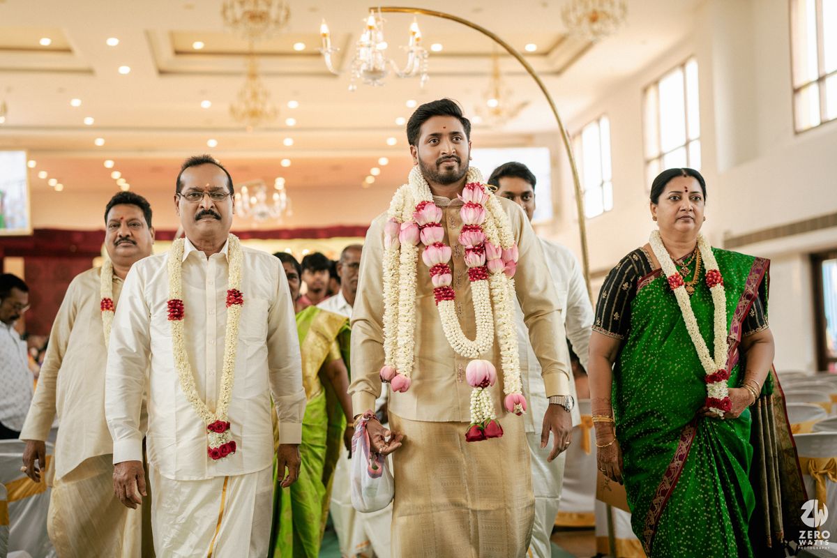 Indian groom in cream sherwani walks with family wearing floral garlands during traditional wedding ceremony.