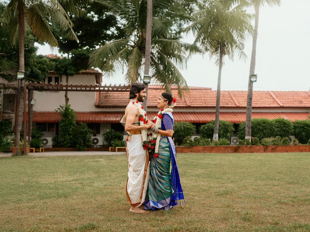 Couple in traditional South Indian attire standing on lush green lawn with palm trees and heritage building backdrop.