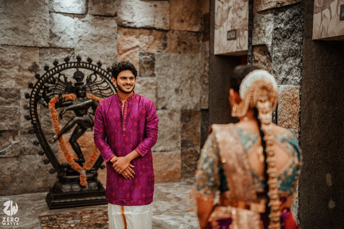 Groom in purple kurta smiles at bride near Nataraja statue in traditional South Indian wedding.