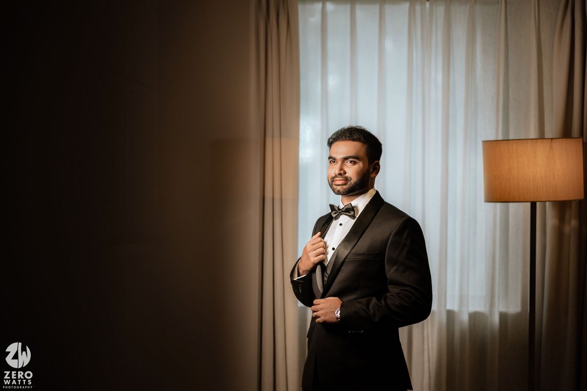 Groom in black tuxedo and bow tie holding microphone, standing by curtained window with warm lamp light.