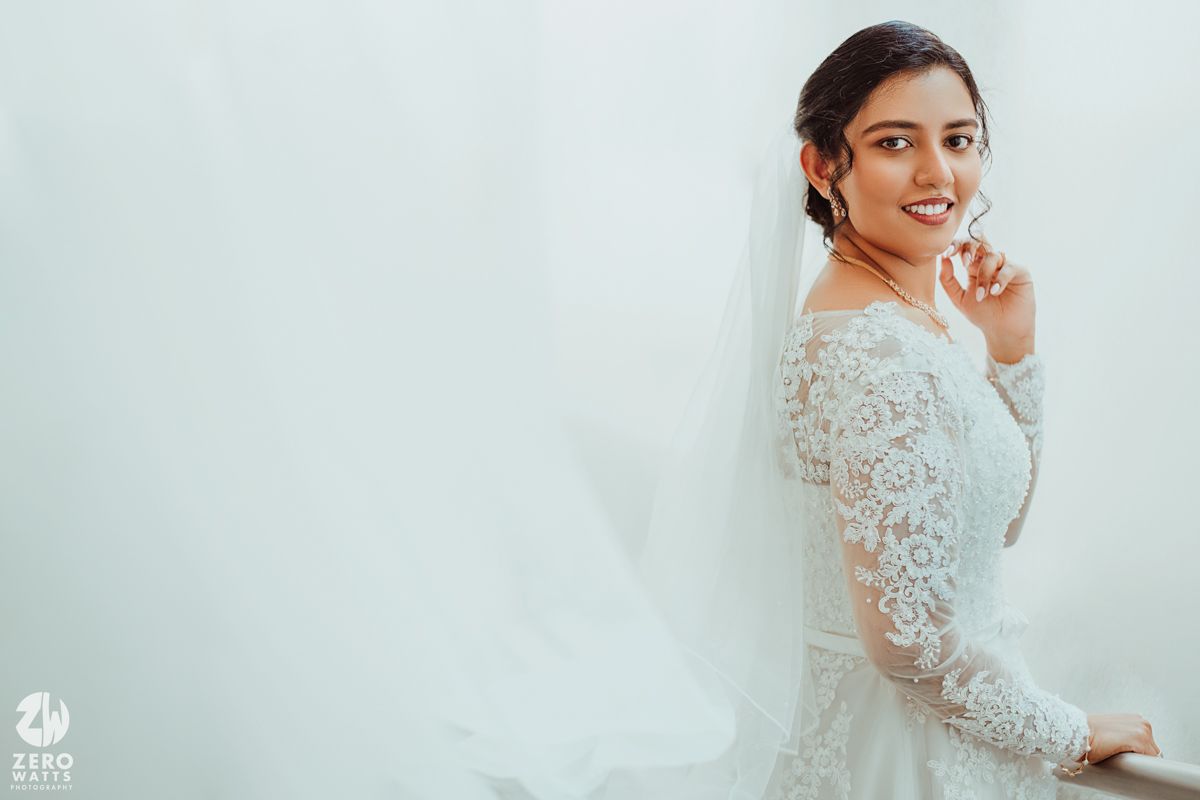 A bride in a long-sleeved white lace wedding gown poses against a light background.