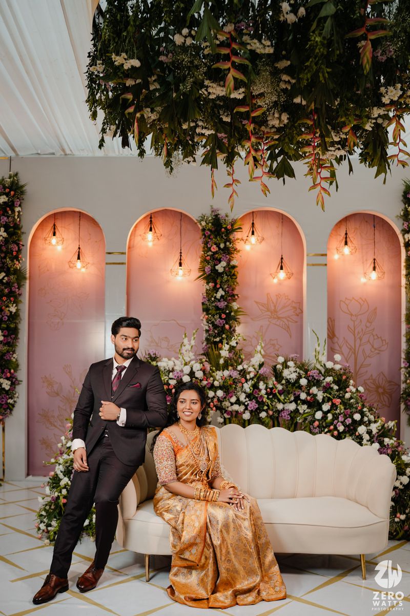 Wedding reception setup with arched backdrop, hanging greenery, and soft pink lighting.