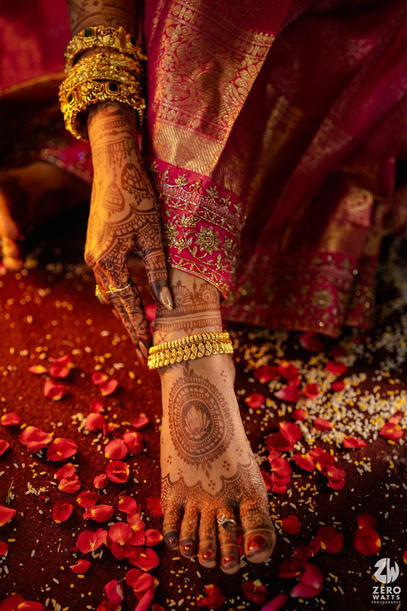 Close-up of Indian bride's henna-adorned hands and feet with gold bangles, anklets, and red rose petals on the floor.
