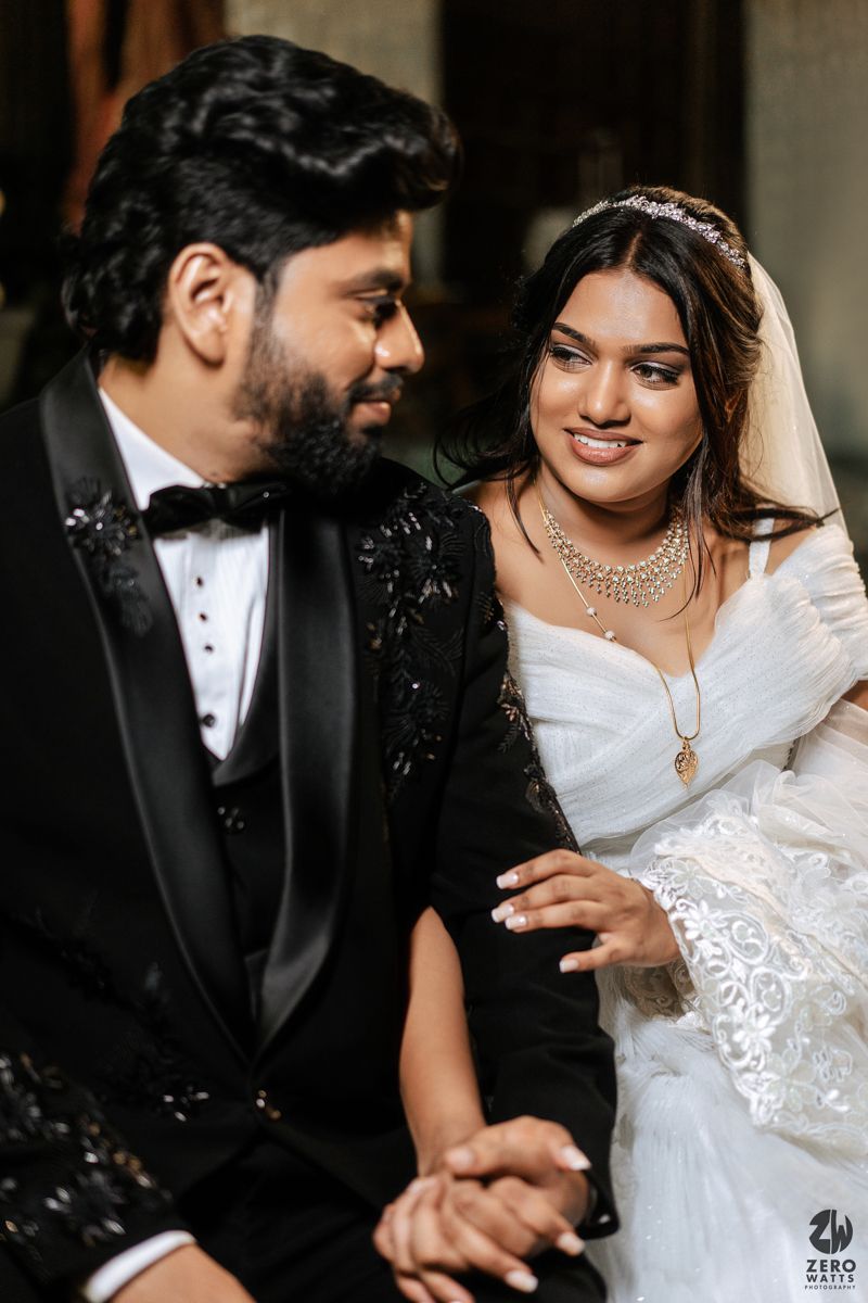 Indian bride in white gown and groom in black tuxedo holding hands and smiling at each other.
