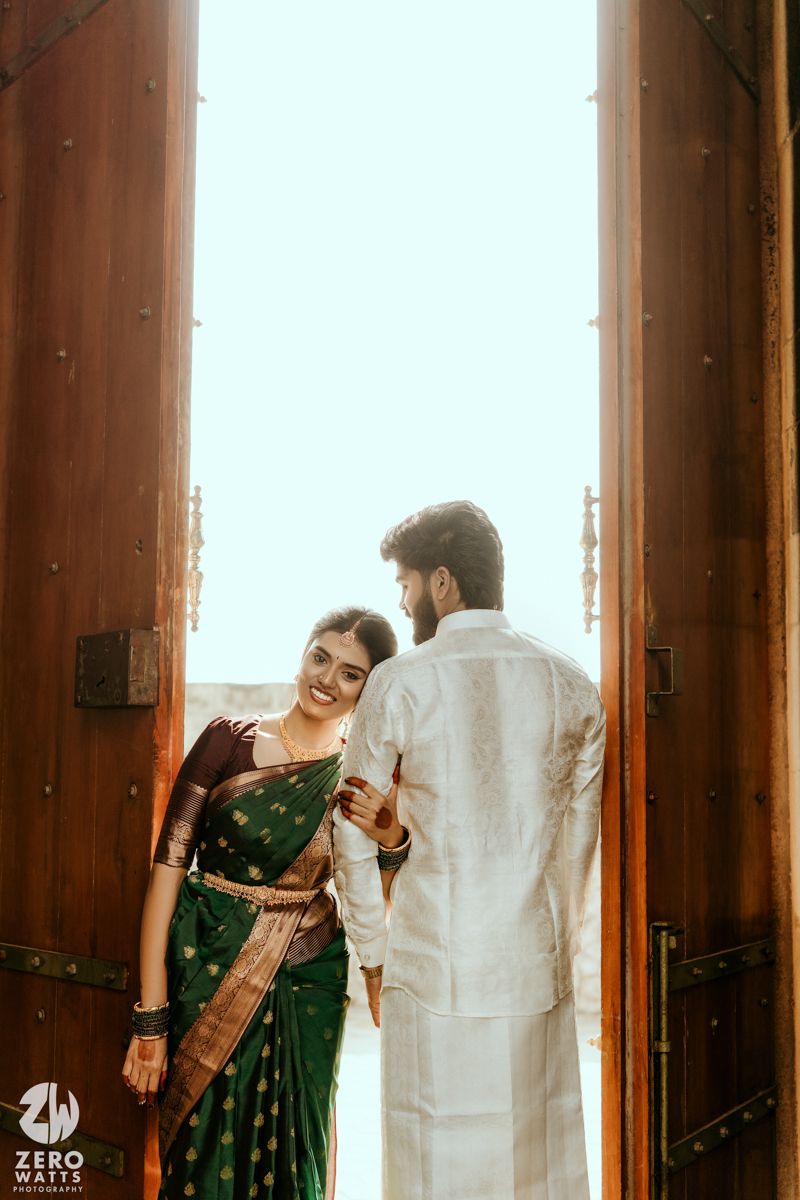 Smiling bride in green silk saree holds groom's hand in white kurta framed by ornate wooden temple doors.