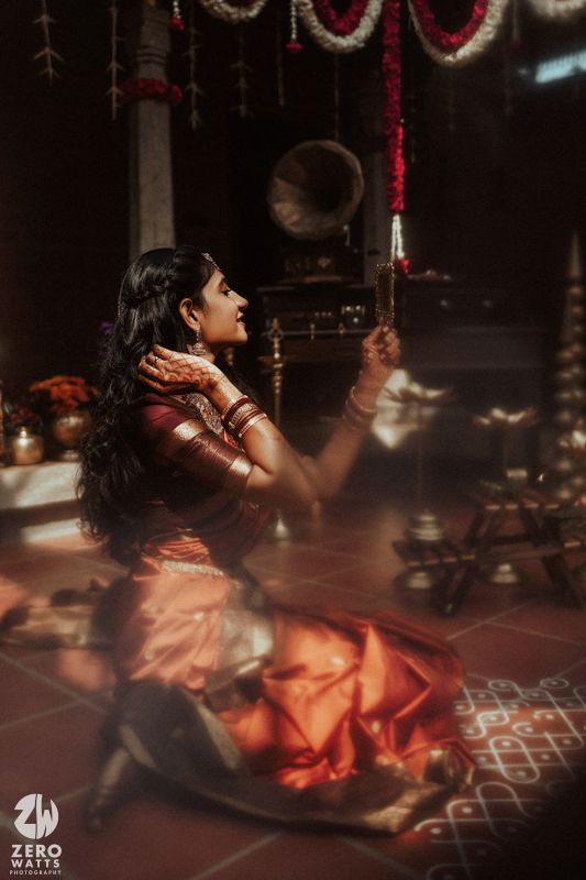 A woman in a red saree sits on the floor holding a mirror, surrounded by traditional Indian decor and kolam.