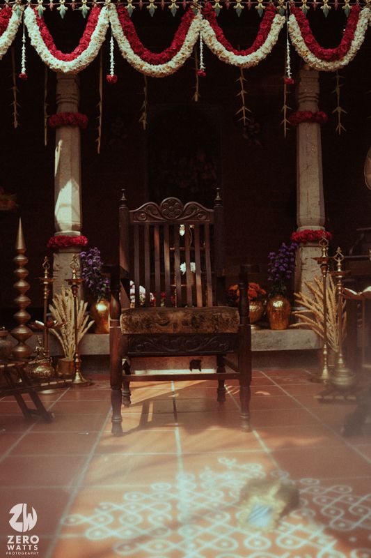 Traditional South Indian wedding mandap with carved wooden chair, brass lamps, and red white flower garlands.