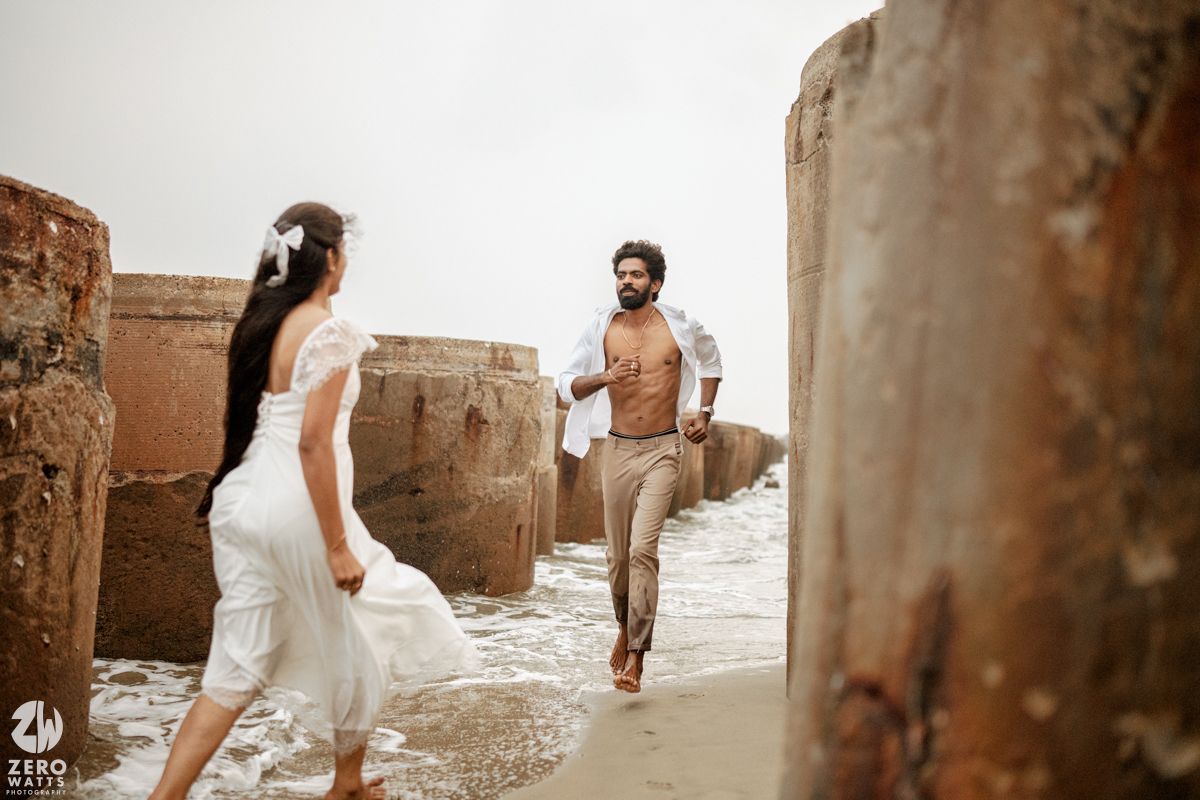 Man in brown shirt running toward woman in white dress between stone pillars on a misty beach shore.