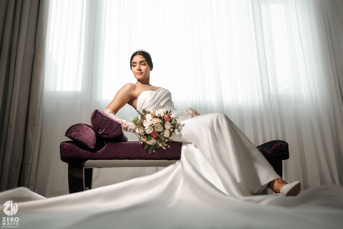 Bride in white wedding dress reclines on purple velvet bench holding a pastel floral bouquet.