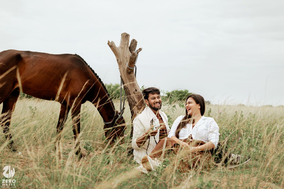A couple poses romantically in a field with horses visible in the distance.