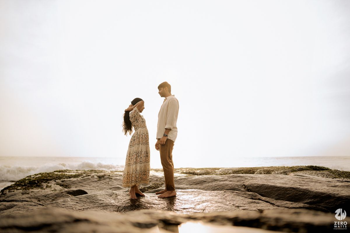Two people share a tender moment on coastal rocks with soft light and ocean views.