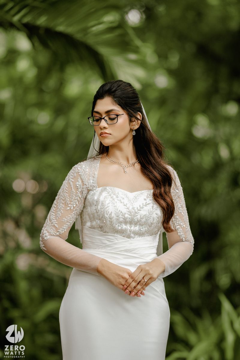 A bride in a white lace wedding dress poses in a garden setting with soft bokeh background lighting.