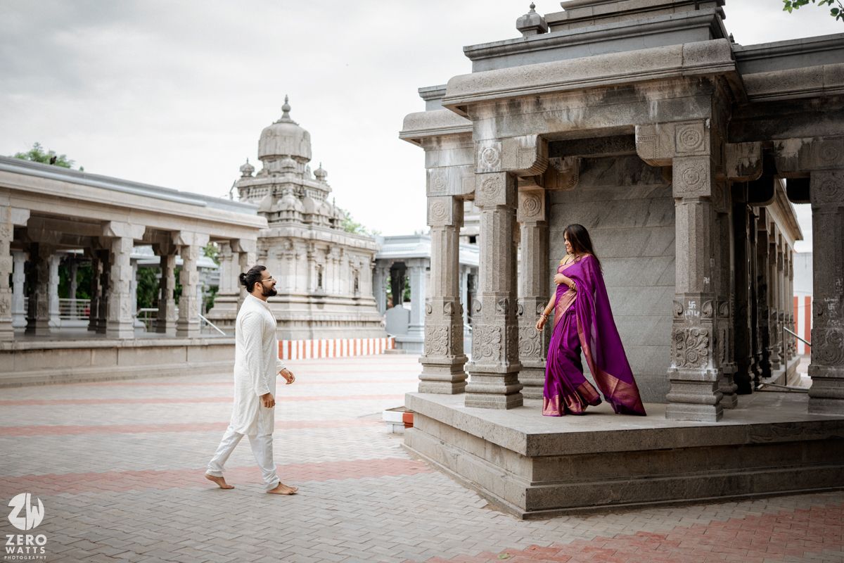 Two people in traditional Indian attire stand in an ornate temple courtyard with white stone pillars and domed architecture.