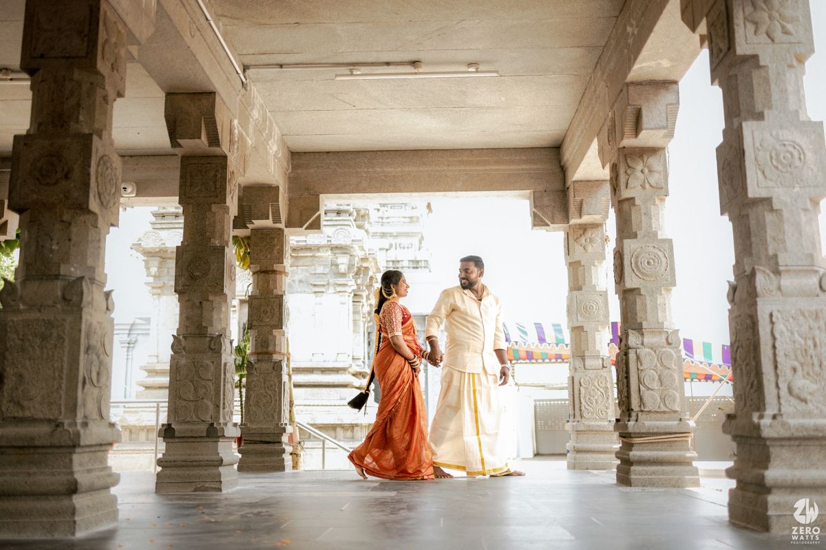 Ancient stone pillars line a corridor in a traditional Indian temple with orange-robed figures visible in the distance.