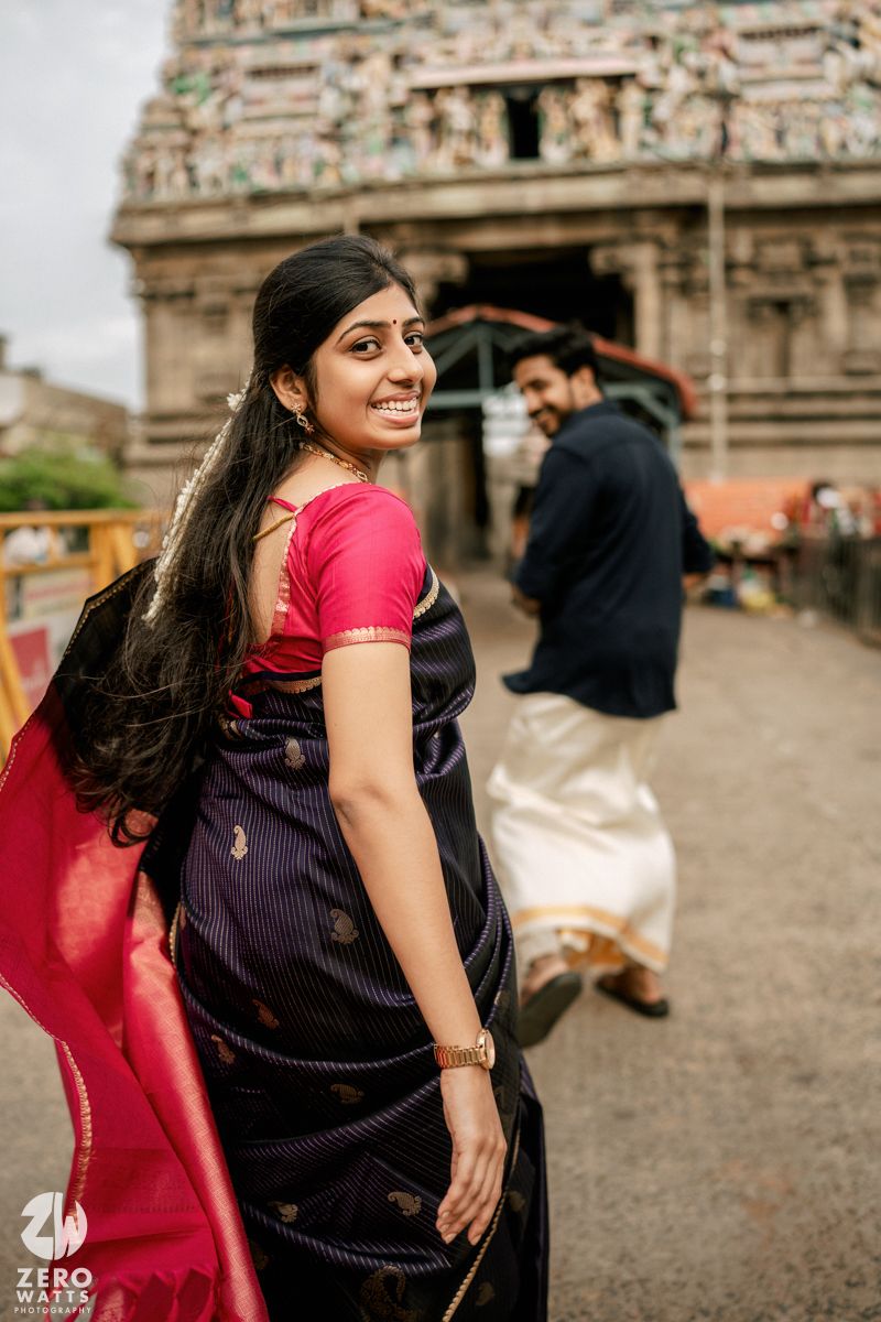 A pair dressed in traditional Indian clothing share a joyful moment outside a temple with detailed carvings.