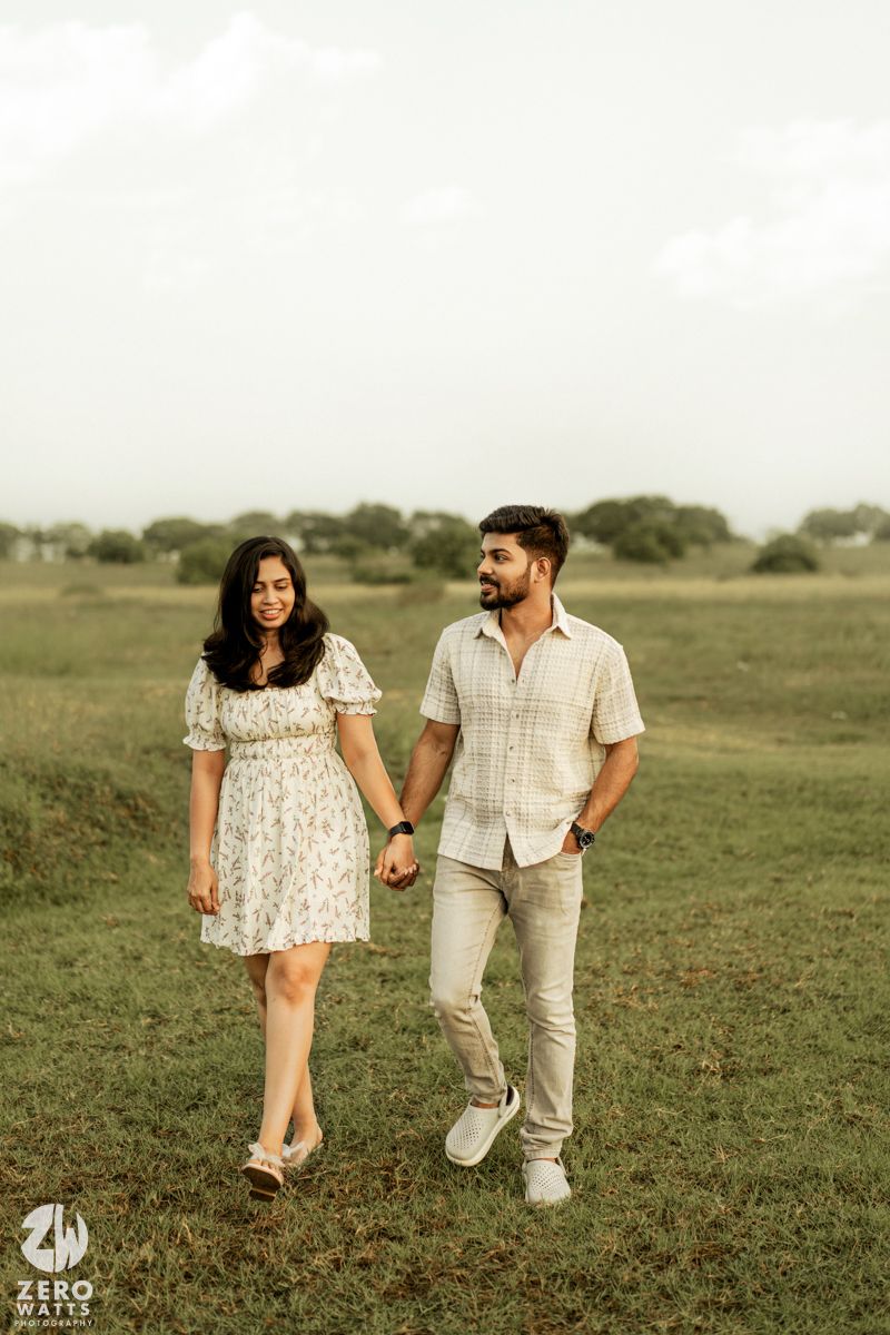 A couple in coordinated white outfits walks hand in hand through a grassy meadow at sunset.